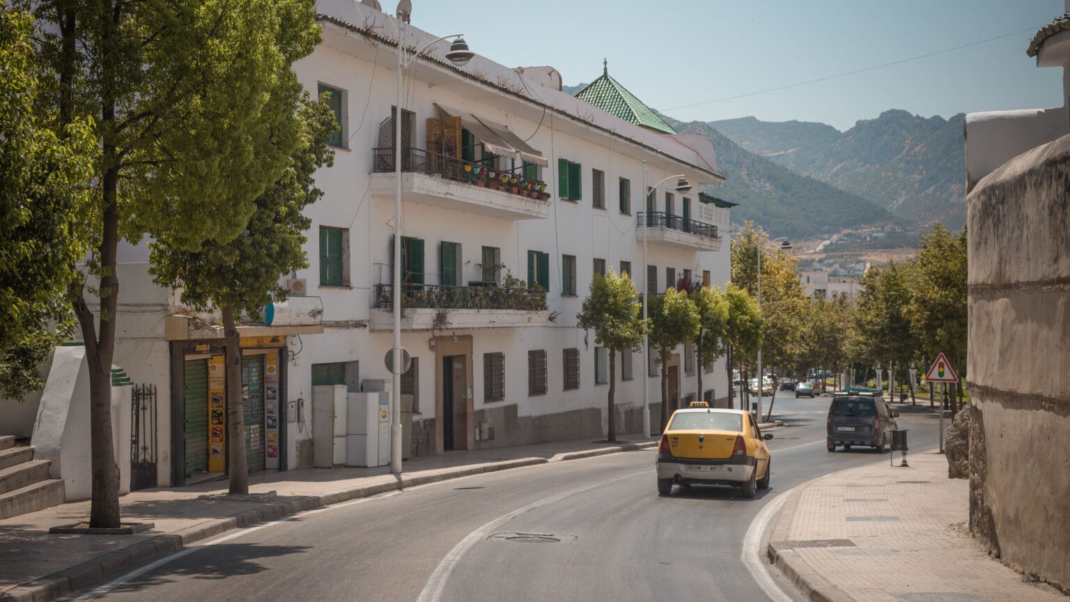 A quiet street in a Rif town: whitewashed buildings with green shutters, a yellow taxi, and mountains in the distance